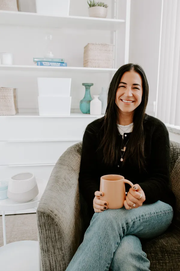 Ellie smiling warmly while sitting in a cozy chair, holding a peach-colored mug